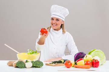 Portrait of cheerful female chef sitting at the table and showing tomato while making healthy meal.