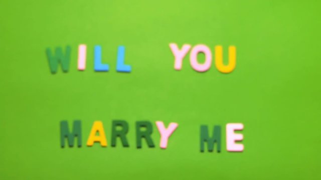 Wood characters of different colors arranged in a word will you marry me on a green background with blur effect