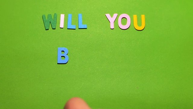 Wood characters of different colors arranged in a word will you be my valentine's on a green background with hand slide
