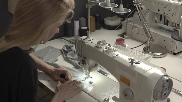 Blonde Caucasian female in black dress sitting at table with machine in sewing classroom stitching grey piece of fabric
