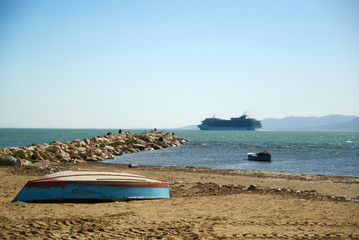 blue, wooden boat, upside down, lying on an empty beach, by the sea, far away in the sea swims a large cruise ship