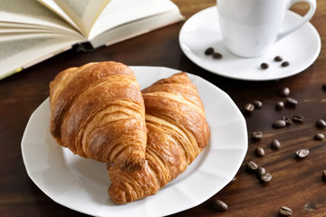 Breakfast scene with fresh croissants, coffee cup, coffee beans and open book. Relaxation morning scene with hot coffee.