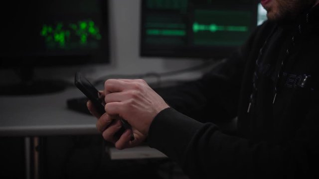 Serious man with brown hair, glasses on the eyes, stubble beard, wearing a black hoodie intently typing on your phone which is located in a dark room, like a secret bunker