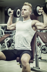 Fototapeta premium Attractive young man training shoulders with dumbbells sitting on bench in gym
