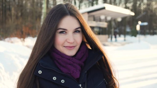 young attractive woman with long dark hair in black coat and knitted scarf look at camera and blow kisses while walking in the snowy park under the sunlight