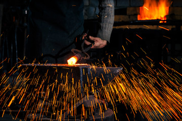 The blacksmith manually forging the molten metal on the anvil in smithy with spark fireworks