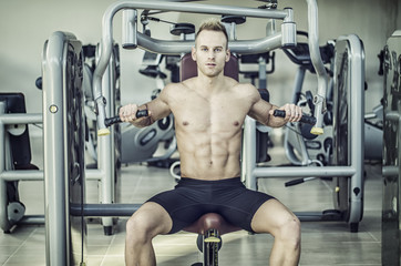 Fototapeta premium Muscular, shirtless young man in gym exercising pecs on pectoral machine