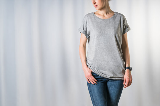 Cropped image. Front view of young woman, dressed in light gray t-shirt and blue jeans, standing on light gray background with his hand in pocket of jeans. Mock up. Space for text, advertising.