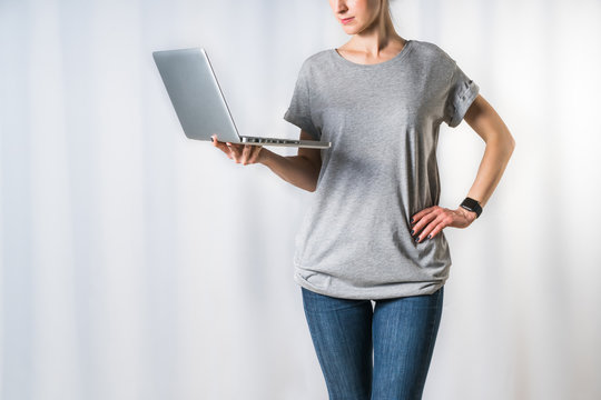Front view of young woman, dressed in gray t-shirt and blue jeans, standing on light gray background and holding laptop. Mock up, space for text, advertising. Girl using digital gadget,checking email.