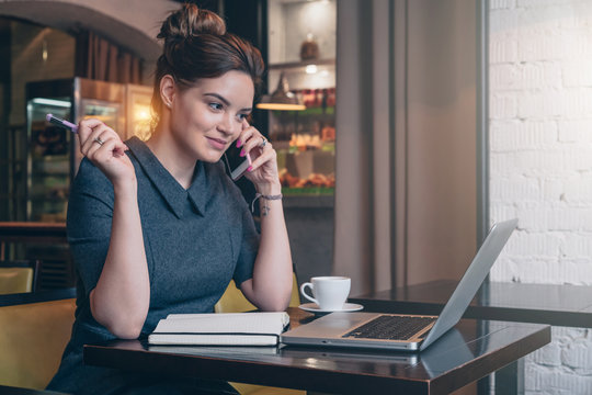 Young business woman in gray dress sitting at table in cafe, talking on cell phone while looking on screen of laptop. On table notebook and cup of coffee. Freelancer working. Student learning online.