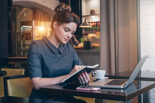 Young businesswoman in gray dress sitting at table in coffee shop and reading book. On table laptop, smartphone and cup of coffee. Student learning online. Freelancer working. Online education.