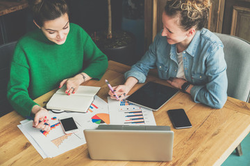 Business meeting. Two smiling young business woman sitting at table and discussing business plan. On table is laptop, smartphone, tablet, paper charts and notebook.Freelancers working online,blogging.