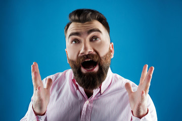 man with a beard in a pink shirt on a blue background shouting emotsianalno hands up  and yelling, screaming