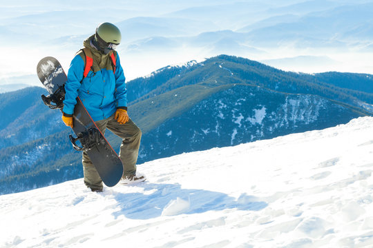 Snowboarder in helmet walking to the very top of a mountain with his snowboard in hand