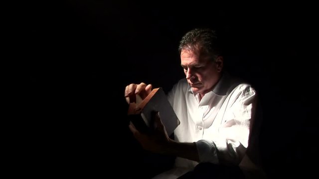 A man spot lighted by a fog filled beam of light  reading a bible and praying.