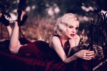 Naklejka premium Piękna i elegancka blondynka z czerwonymi ustami i falami włosów na sobie czerwoną koszulę nocną z winem, pozowanie na łóżku na zewnątrz jesienią, styl retro vintage i moda. Wyretuszowane ujędrnienie