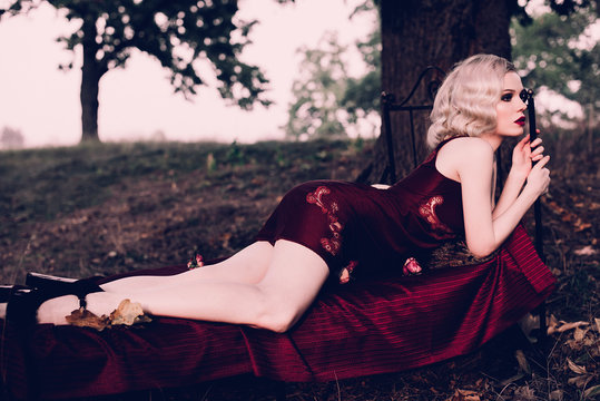 Beautiful and elegant blonde woman with red lips and hair waves wearing wine red nightie posing on the bed outdoors autumn, retro vintage style and fashion. Retouched toning shot