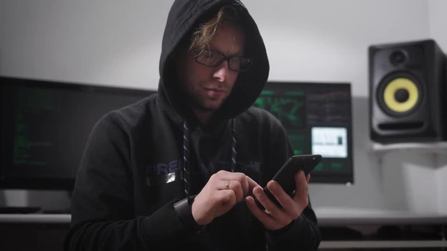 Strained man with hooded jackets by color, with black-rimmed glasses on his eyes, brown hair and stubble beard sitting n the chair and the stress is typing a message on the touch screen