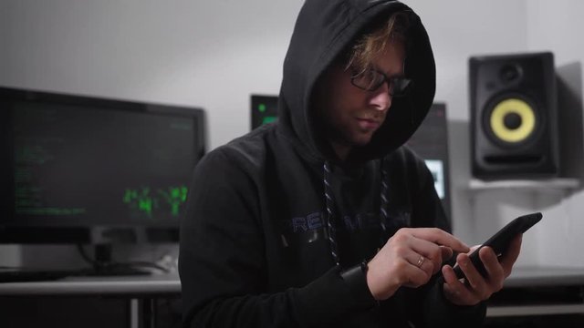 The underground organization. Laptop on the monitors started cracking process. A hacker with a smartphone on a hand sits on a chair. The guy scans and enters the information in the smartphone.