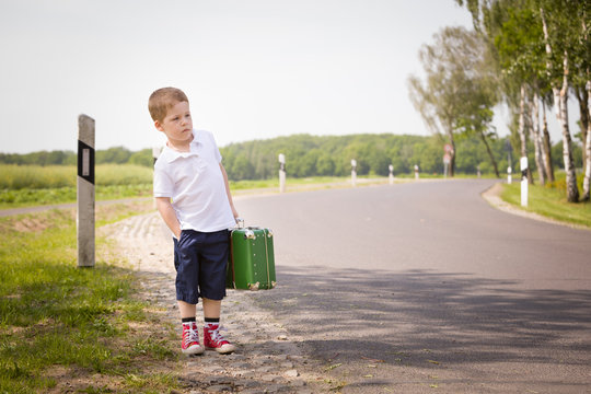 Adorable little kid boy in a white polo shirt and shorts holds a green retro suitcase and  stands on the countryside road waiting for a bus or a car. Ready for travel. Vacation time. Child on the road