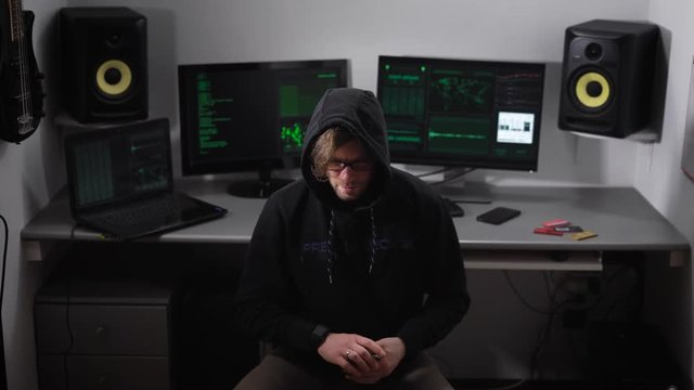 Hooded hacker in black clothes sitting on a chair in the room and counting prepayment for hacking network. After checking money man puts it into the pocket and turns to computer to make cyber crime.