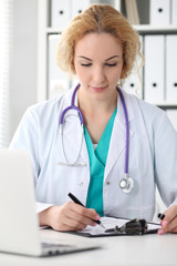 Happy blonde female doctor sitting at the table and filling up medical history form. Medicine, healthcare and help concept. Physician ready to examine patient