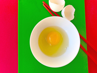 Raw egg in white bowl with red chopsticks and eggshells on green and red cutting boards