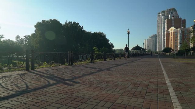 A sporty young man slowly runs along the sidewalk from the back. The male wears shorts, a tank top, sneakers and a runner's armband. Beautiful city scale is in the background.
