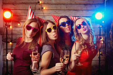 Birthday party girlfriends. Beautiful girls in caps, and colored glasses with champagne glasses grimace shows language.
