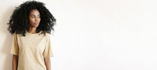 People and lifestyle. Studio shot of gorgeous African girl with Afro hairstyle wearing oversize t-shirt standing isolated at white wall and looking away with serious thoughtful expression on her face