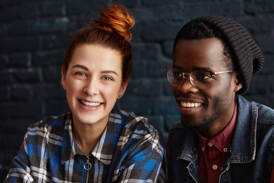 Beautiful redhead student girl with charming smile spending nice time with her handsome stylish African groupmate wearing black hat and glasses. Interracial couple in love having fun at coffee shop