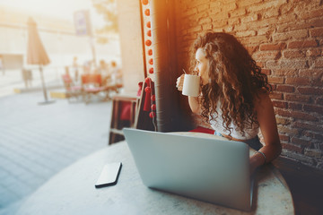 Beautiful curly brunette girl drinks tea or coffee while sitting with portable laptop in street...