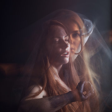 Beautiful model with red hair posing behind a black fabric and a veil in a studio