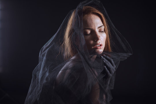 Beautiful model with red hair posing behind a black fabric and a veil in a studio