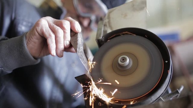Old man sharpening his ax on a grinder in an workshop