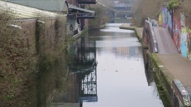 A Tilting Shot from the murky rippling waters of a Birmingham canal to show the weedy pathway, graffiti walls and industrial urban buildings.
