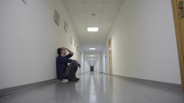 Young man squats leaning to wall in long corridor and waiting