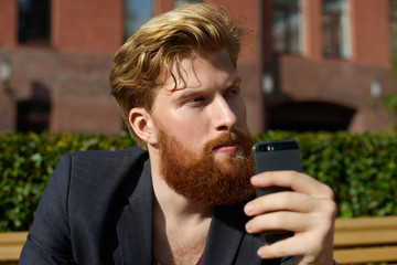 Handsome blond hipster dream outdoor with phone in hand. Thinking guy sitting on the bench in the street with old red brick buildings concept.