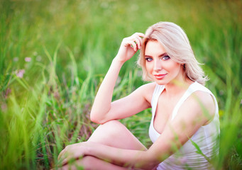 Naklejka na ściany i meble Smiling beautiful young girl sitting among green grass and flowers