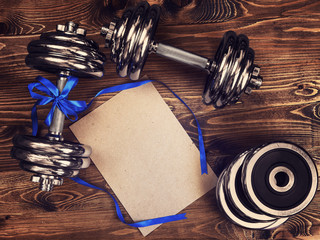 Toned image of metal dumbbells, blue atlas ribbon and a sheet of craft paper on a wooden background - 136733573