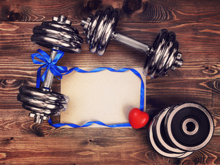 Toned image of metal dumbbells, blue atlas ribbon, red heart and a sheet of craft paper on a wooden background - 136733707