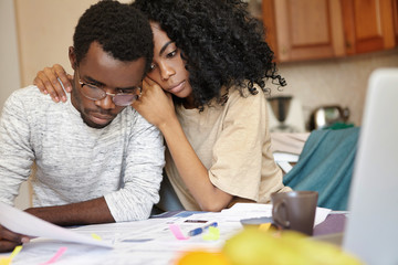 African couple is bankrupt. Sad male in glasses holding piece of paper feeling effortless to do something to save his business while his strong wife hugging him tenderly, trying to cheer him up
