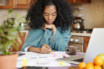 Indoor shot of beautiful young African housewife holding pencil, filling in papers while managing...
