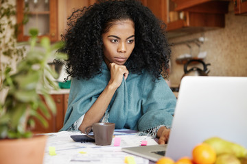 Beautiful African woman sitting in front of laptop computer, looking at screen with serious and...