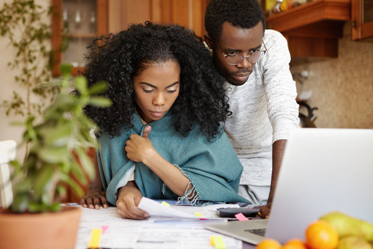 Young dark-skinned couple managing domestic budget and paying bills using online banking app on generic laptop. Wife with piece of paper calculating family expenses together with her husband