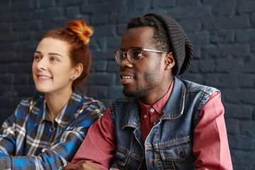 Friendhip and people concept. Beautiful young redhead Caucasian female with hair bun relaxing at coffee shop with modern interior together with her attractive dark-skinned boyfriend in trendy clothes