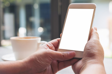 Mockup image of hands holding white mobile phone with blank white screen and hot coffee cup in vintage cafe