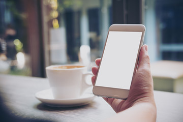 Mockup image of hands holding white mobile phone with blank white screen and hot coffee cup in vintage cafe