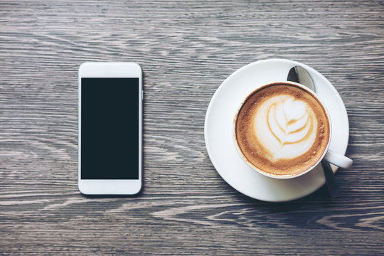 Mockup image of white mobile phone with blank black screen and hot latte coffee on vintage wood table in cafe