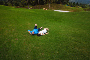 Fototapeta na wymiar Young beautiful couple holding hands, lying at green grass of golf course in summer. man wear the blue shirt and the girl in a white dress. Concept of honeymoon